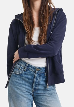 Person wearing a navy zip-up hoodie over a white tank top and faded blue jeans, with one arm crossed over the waist.