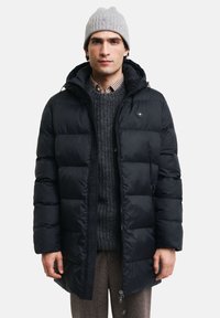 Sort puffjakke med hætte, tykt quiltet design, frontlynlås og mærke på brystet, båret over en grå sweater og ternet skjorte.