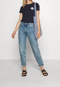 Námořnické bavlněné tričko s grafikou mušle a textem, světle modré džíny s vysokým pasem a černé sandály. Taška je pruhovaná modro-bílá.