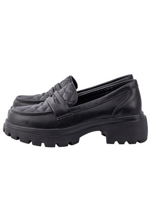Personne portant un pantalon ample large texturé rose, un top court noir et des mocassins noirs épais, posant avec une jambe levée.