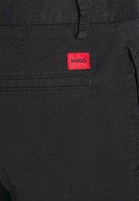 Gros plan de la poche d'un pantalon noir avec une étiquette rectangulaire rouge affichant le nom de la marque "HUGO" en lettres noires.