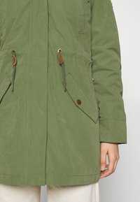 Manteau vert mi-long avec cordons et grandes poches à rabat avec fermetures par boutons, porté sur un pantalon blanc, montrant le bras et la main droite.