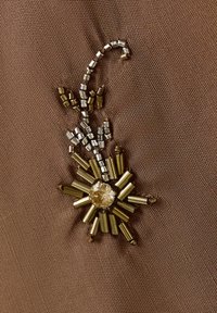 Broderie de perles dorées et argentées en forme florale avec une pierre centrale sur fond de tissu marron texturé.