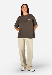 Hnedé bavlnené voľné tričko s krátkymi rukávmi a malým bielym logom na ľavej hrudi, v kombinácii so svetlými textúrovanými nohavicami a teniskami.
