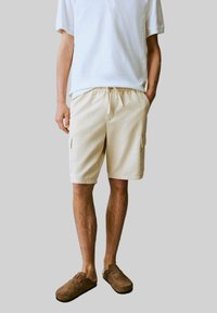 Beige cargoshorts med sidofickor, tillverkade av lättviktig tyg. Utrustad med dragsko i midjan och avslappnad passform, kombinerad med en vit pikétröja.