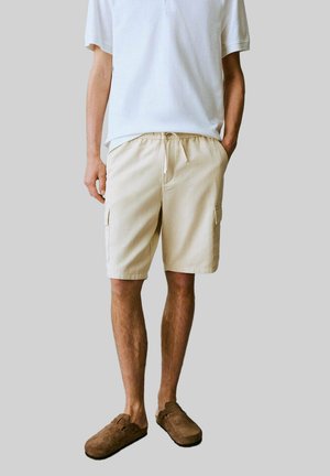 Beige cargoshorts med sidofickor, tillverkade av lättviktig tyg. Utrustad med dragsko i midjan och avslappnad passform, kombinerad med en vit pikétröja.