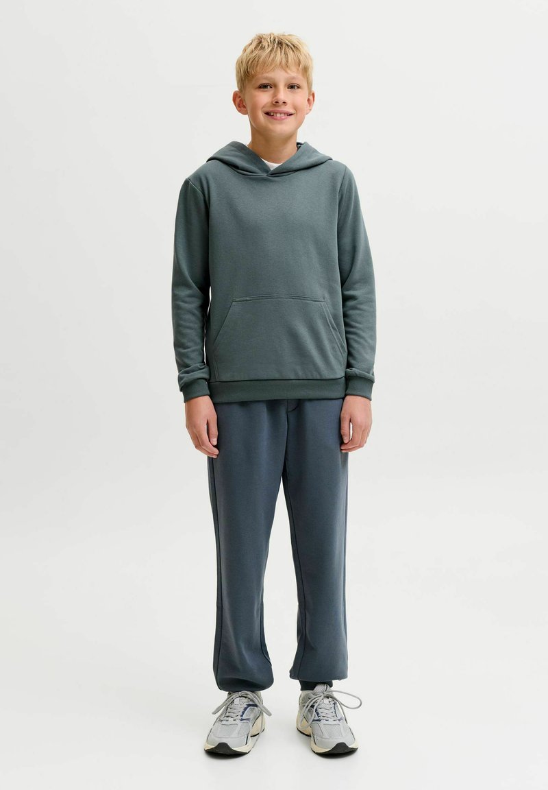 Garçon blond debout et souriant, portant un sweat à capuche vert foncé, un pantalon sombre et des baskets blanches sur un fond clair uni.