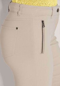 Beige Hose mit glatter Oberfläche, ausgestattet mit einer seitlichen Reißverschlusstasche und einer normalen Gesäßtasche mit einem Metallknopf.