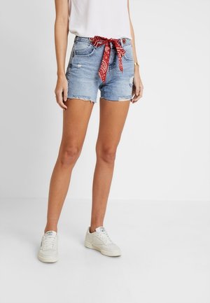 Kobieta nosząca jasnoniebieskie postrzępione szorty jeansowe z czerwonym paskiem bandana, białe trampki i biały top bez rękawów, stojąca na białej podłodze.