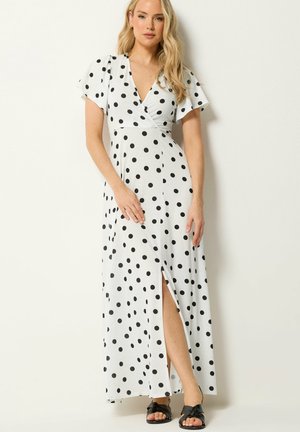 Vrouw draagt een witte wikkeljurk met zwarte stippen en een voorzijde met een splitsing, gecombineerd met zwarte slippers, staand tegen een witte muur.