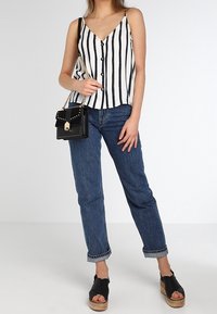 Biała i czarna w paski bezrękawnik z guzikami, w połączeniu z niebieskimi, podwiniętymi dżinsami i czarnymi sandałami na platformie, trzymająca małą czarną torebkę.