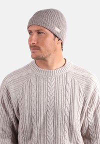 Bonnet gris côtelé en tricot avec une étiquette, associé à un pull gris clair en maille torsadée avec des motifs texturés et un col rond.