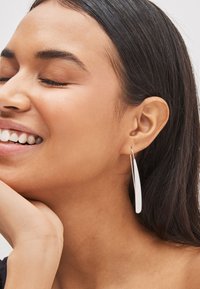 Boucles d'oreilles pendantes allongées en argent, à surface lisse et polie, suspendues à un fil fin, présentées sur un modèle aux cheveux foncés.