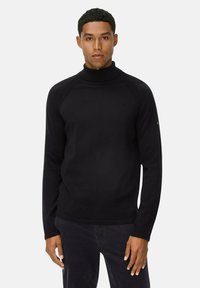 Mann in schwarzem Rollkragenpullover und dunkler Hose, der vor einem schlichten hellen Hintergrund steht und nach vorne blickt.