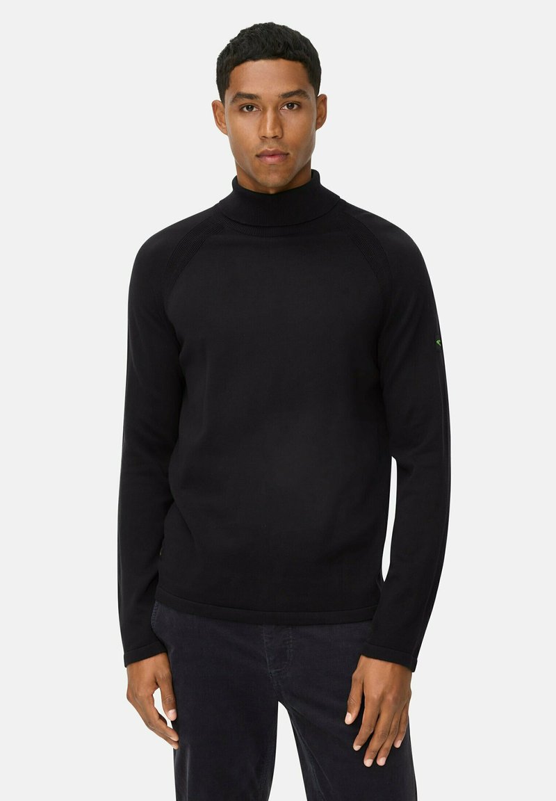 Mann in schwarzem Rollkragenpullover und dunkler Hose, der vor einem schlichten hellen Hintergrund steht und nach vorne blickt.