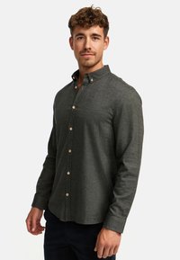 Chemise boutonnée vert foncé en tissu texturé avec un motif vertical et des boutons contrastants de couleur claire. Elle présente un col classique et des manches longues.