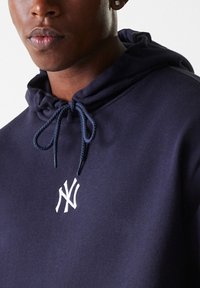 Marineblauer Hoodie mit einer Kordelzugkapuze, ausgestattet mit einem weißen gestickten Logo auf der Brust. Hergestellt aus einem weichen Stoff mit glatter Textur.