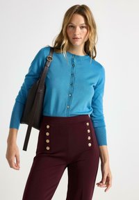 Cardigan button-up azzurro chiaro con maniche lunghe, abbinato a pantaloni bordeaux a vita alta con dettagli di bottoni dorati sulle tasche.