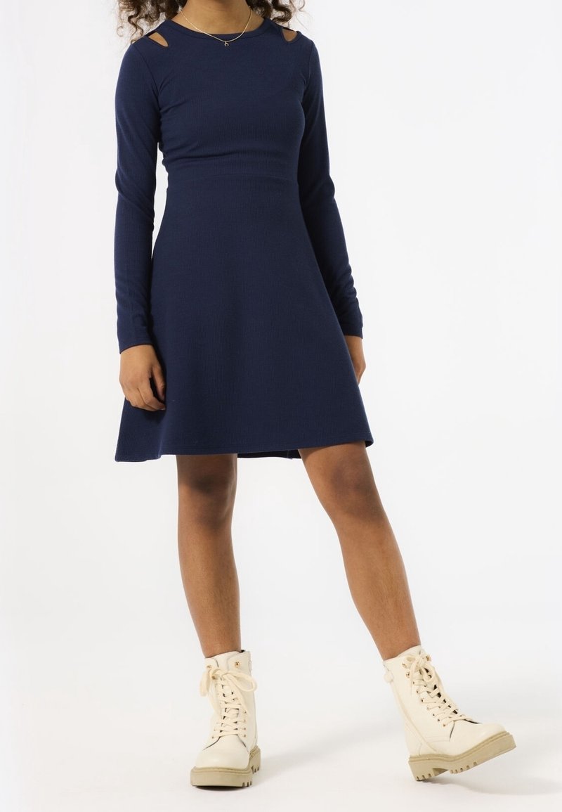 Jeune personne portant une robe bleu marine à manches longues avec des découpes aux épaules et des bottes crème à lacets, debout sur un fond blanc.