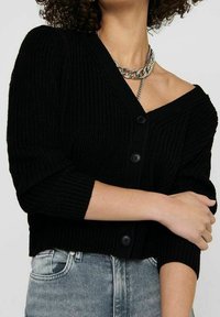 Cardigan nero a maglia con scollatura a V, maniche a tre quarti e chiusura con bottoni. Indossato su un outfit in denim chiaro. Collana a catena come accento.