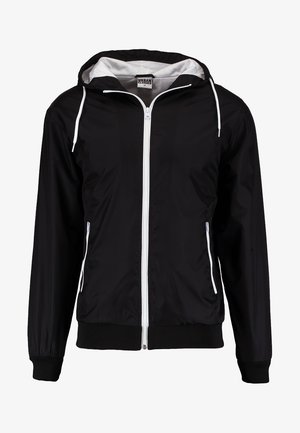 Zwarte windbreakerjas met capuchon, witte rits en zijzakken. Gemaakt van lichtgewicht materiaal met geribbelde manchetten en zoom.