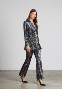 Traje de terciopelo en gris oscuro con un patrón veteado. Presenta un blazer ajustado y pantalones de pierna recta, complementados con tacones altos negros.