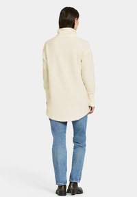 Cremefarvet fleece-pullover med høj krave, lange ærmer og rundet kant, parret med blå jeans og mørke støvler.