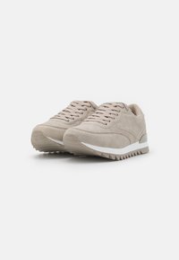 Sapatilhas bege com upper texturizado, apresentando fecho com atacadores e um entral do pé branco. A sola exterior tem um design estriado para tração.