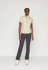 Beżowa kamizelka bez rękawów puchowa nałożona na jasnobeżową koszulę, w zestawieniu z ciemnoszarymi spodniami. Białe sneakersy i okulary przeciwsłoneczne dopełniają strój.