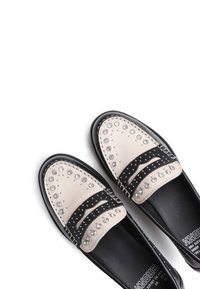 Mocasines de cuero en negro y crema con tachuelas plateadas, bandas horizontales en contraste, punta redondeada y detalles cosidos a lo largo de la suela.