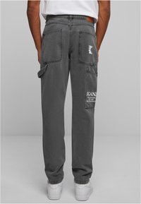 Šedé bavlněné cargo kalhoty mají více kapes, kontrastní šití a koženou nášivku na pase. Na pravé noze je viditelný bílý text.
