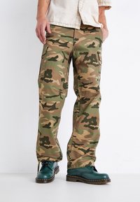 Camouflage cargobroek in groen, bruin en tan met een losse pasvorm, voorzien van meerdere zakken, gecombineerd met groene veterschoenen.