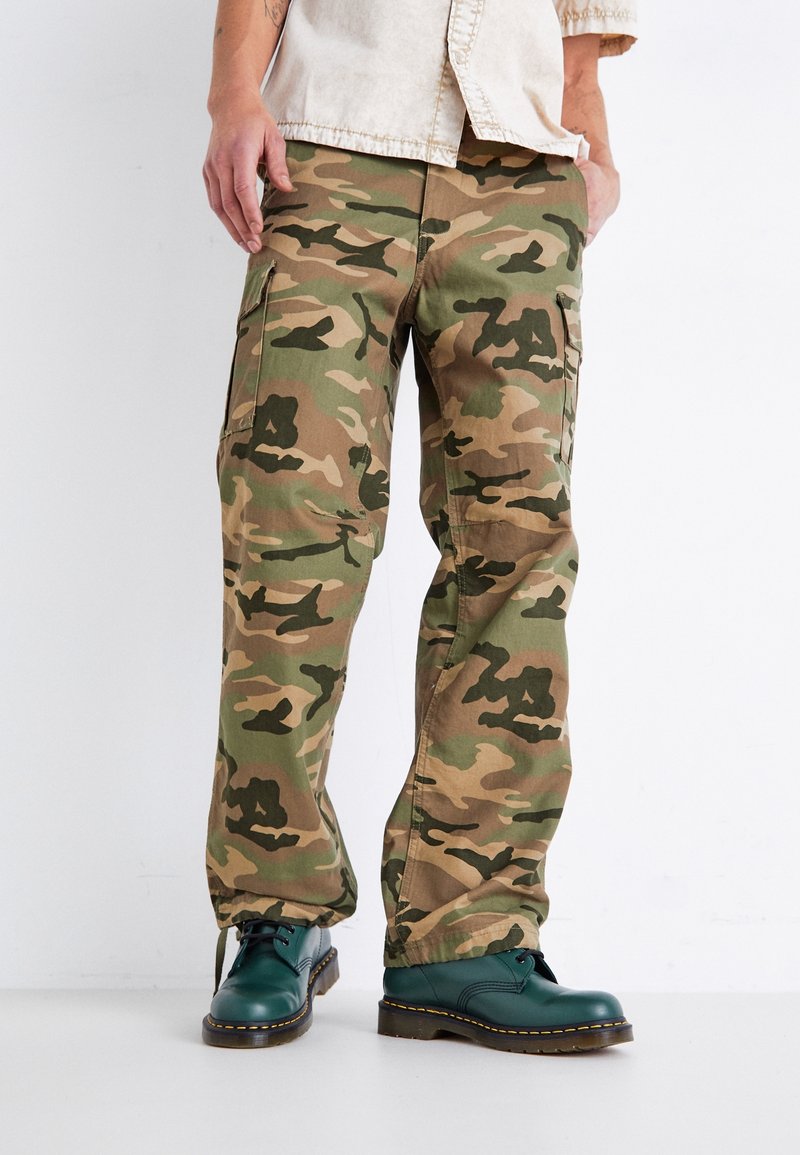 Camouflage cargobroek in groen, bruin en tan met een losse pasvorm, voorzien van meerdere zakken, gecombineerd met groene veterschoenen.