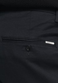 Pantalones de tela negra con un diseño elegante, bolsillo con cremallera y un botón negro. La etiqueta "GANT" es visible en el bolsillo.