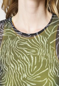 Διαφανής μπλούζα με πράσινο φόντο και λευκούς και μαύρους αφηρημένους κειμενικούς σχεδιασμούς. Στρογγυλή λαιμόκοψη με αντίθετη μαύρη σκελέα.