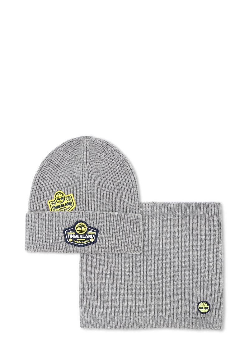Ensemble bonnet et écharpe en tricot gris, avec une texture côtelée et des logos Timberland en jaune et bleu. Les accents incluent des étiquettes de marque.