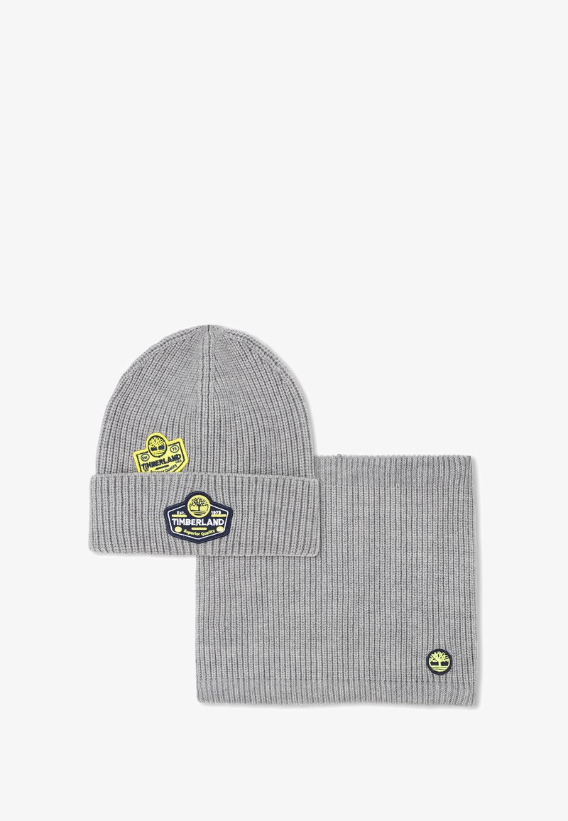 Ensemble bonnet et écharpe en tricot gris, avec une texture côtelée et des logos Timberland en jaune et bleu. Les accents incluent des étiquettes de marque.