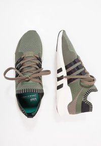 Gröna och svarta sportskor med en texturerad ovandel, snörning, vadderad sula och framträdande logotypdetaljer på tungan och sidorna.