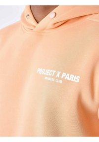Sudadera de color melocotón con una textura suave, que presenta un logo en blanco: "PROJECT X PARIS MEMBERS' CLUB" en el pecho y un cuello con botones a presión.