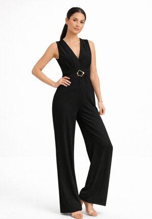 Kobieta o ciemnych włosach, ubrana w czarny bezrękawnikowy kombinezon ze złotą klamrą, stojąca z jedną ręką na biodrze i beżowymi szpilkami na białym tle.