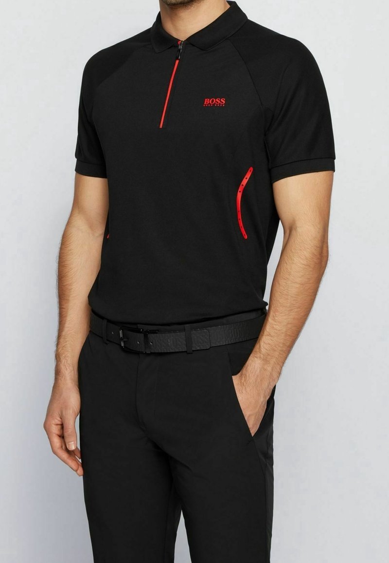 Homme portant un polo noir à manches courtes avec une fermeture éclair et des accents rouges, une ceinture noire et un pantalon noir, debout avec une main dans la poche.