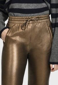 Bronzové metalické joggingové kalhoty s elastickým pasem a stahovací šňůrkou, s bočními kapsami a hladkou, lesklou texturou.