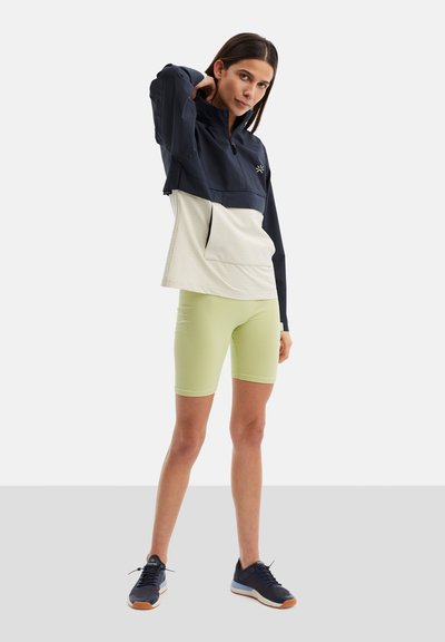 Marinblå och krämfärgad dra-på idrottsjacka med elastisk nederkant, kombinerad med strukturerade ljusgröna cykelshorts och mörka sneakers.