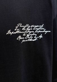 Biely kurzívny text sponzorskej správy vytlačený na čiernej látke, pravdepodobne na hrudnej časti odevu.