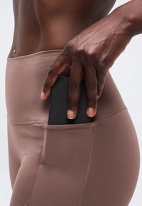 Main glissant un smartphone noir dans la poche latérale d’un legging marron moulant, au niveau de la hanche et de la taille d’une personne à la peau foncée.