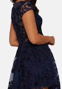 Navy blue dress with short cap sleeves, floral lace embroidery, sheer back, and flared skirt. Textured fabric with a fitted bodice.
