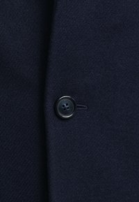 Ciemno granatowa tkanina z pojedynczym czarnym guzikiem cztero-otworowym, na którym wokół krawędzi znajduje się napis "GANT 1949".