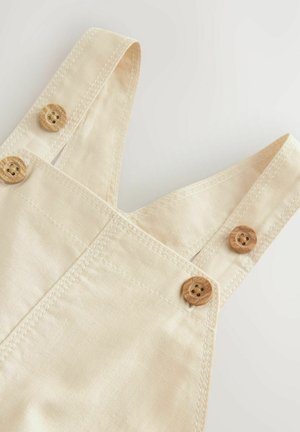 Bežna bombažna kombinezona z nastavljivimi naramnicami, lesenimi gumbi in kontrastnim šivanjem. Gladka tekstura, minimalistična zasnova, brez vzorcev.