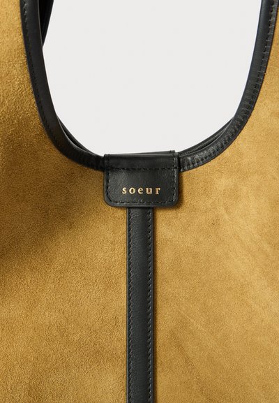 Smeđa suknjasta torba s crnim kožnim detaljima. Sadrži središnju crnu traku s oznakom s logotipom koja prikazuje "soeur" u zlatnim slovima.