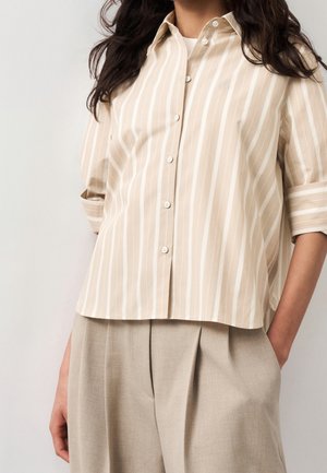 Femme portant une chemise beige et blanche à rayures verticales, boutonnée, avec les manches retroussées, et un pantalon beige plissé, les mains dans les poches.